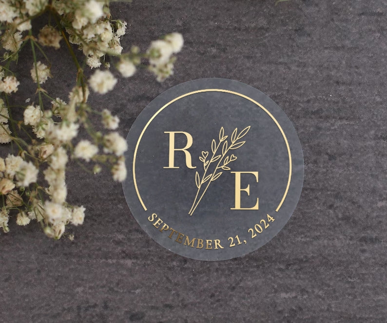 Transparent wedding sticker with golden initials and date, featuring an elegant botanical design