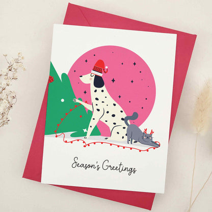 Unleash the holiday spirit with our adorable 'Season's Greetings' card, featuring a festive spotted dog and a cheerful companion decorating a Christmas tree under a starry night sky, perfect for dog lovers and fans of playful designs.