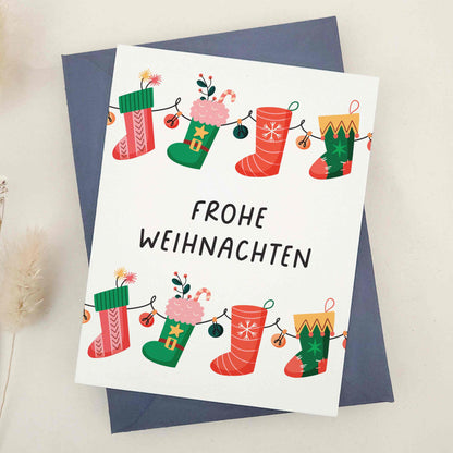 Delightful 'Frohe Weihnachten' card adorned with whimsical Christmas stockings, each festively decorated to capture the cozy and cheerful spirit of the season. The stockings, strung together like a garland, symbolize the connection and joy of the holidays. This card combines traditional German holiday charm with a touch of whimsy, making it perfect for sharing festive wishes.