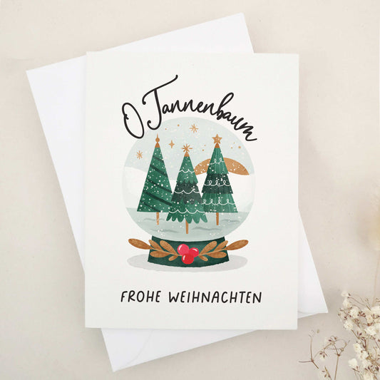 Enchanting 'O Tannenbaum, Frohe Weihnachten' card, paying homage to the classic German Christmas carol. Features a serene snow globe scene with evergreen trees, capturing the essence of 'O Tannenbaum'. The imagery creates a magical, peaceful winter evening atmosphere, reflecting the enduring charm and warmth of the holiday season.