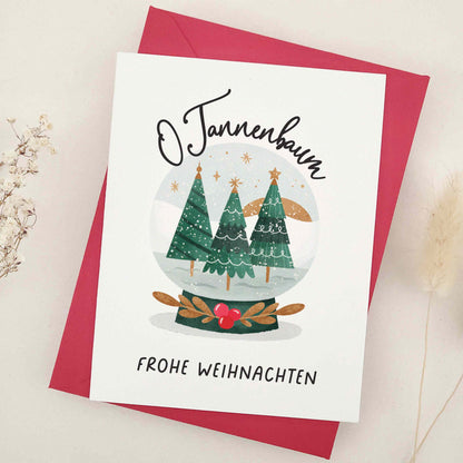 Enchanting 'O Tannenbaum, Frohe Weihnachten' card, paying homage to the classic German Christmas carol. Features a serene snow globe scene with evergreen trees, capturing the essence of 'O Tannenbaum'. The imagery creates a magical, peaceful winter evening atmosphere, reflecting the enduring charm and warmth of the holiday season.