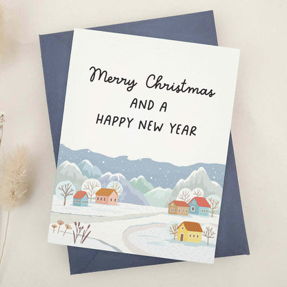 Picturesque 'Merry Christmas and a Happy New Year' card, featuring a tranquil village scene with snow-capped mountains in the background. This beautifully crafted card harmoniously blends nature's grandeur and quaint village charm, symbolizing peace and hope of the festive season. It artistically represents the serene beauty of winter and the joyful anticipation of a new year.