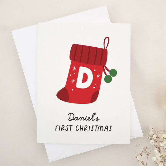 Daniel's First Christmas card: capture the magic of a memorable milestone with a sweet keepsake featuring a cheerful red stocking, bold initial, and festive snowflakes, celebrating a child's first holiday season.