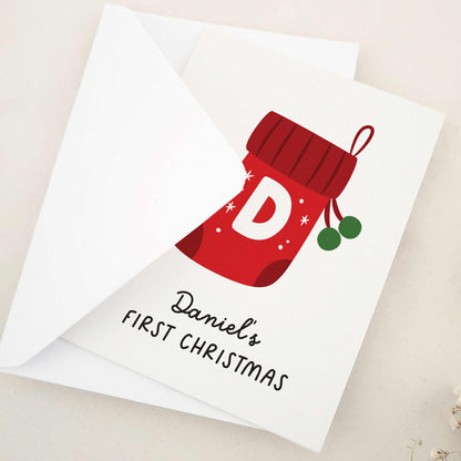 Daniel's First Christmas card: capture the magic of a memorable milestone with a sweet keepsake featuring a cheerful red stocking, bold initial, and festive snowflakes, celebrating a child's first holiday season.