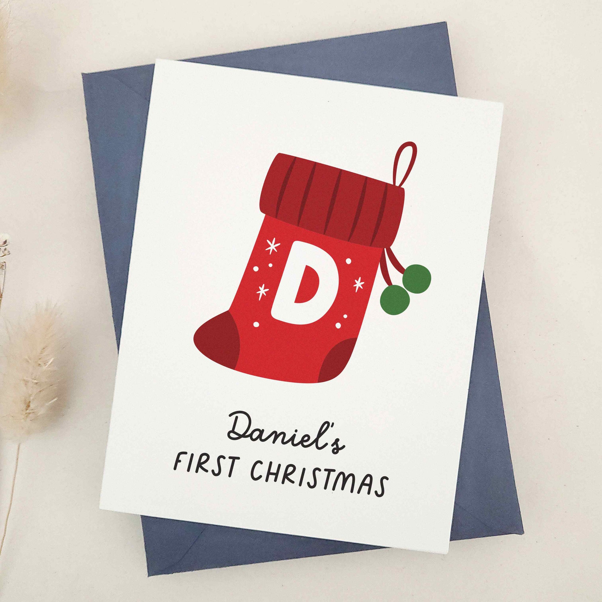 Daniel's First Christmas card: capture the magic of a memorable milestone with a sweet keepsake featuring a cheerful red stocking, bold initial, and festive snowflakes, celebrating a child's first holiday season.