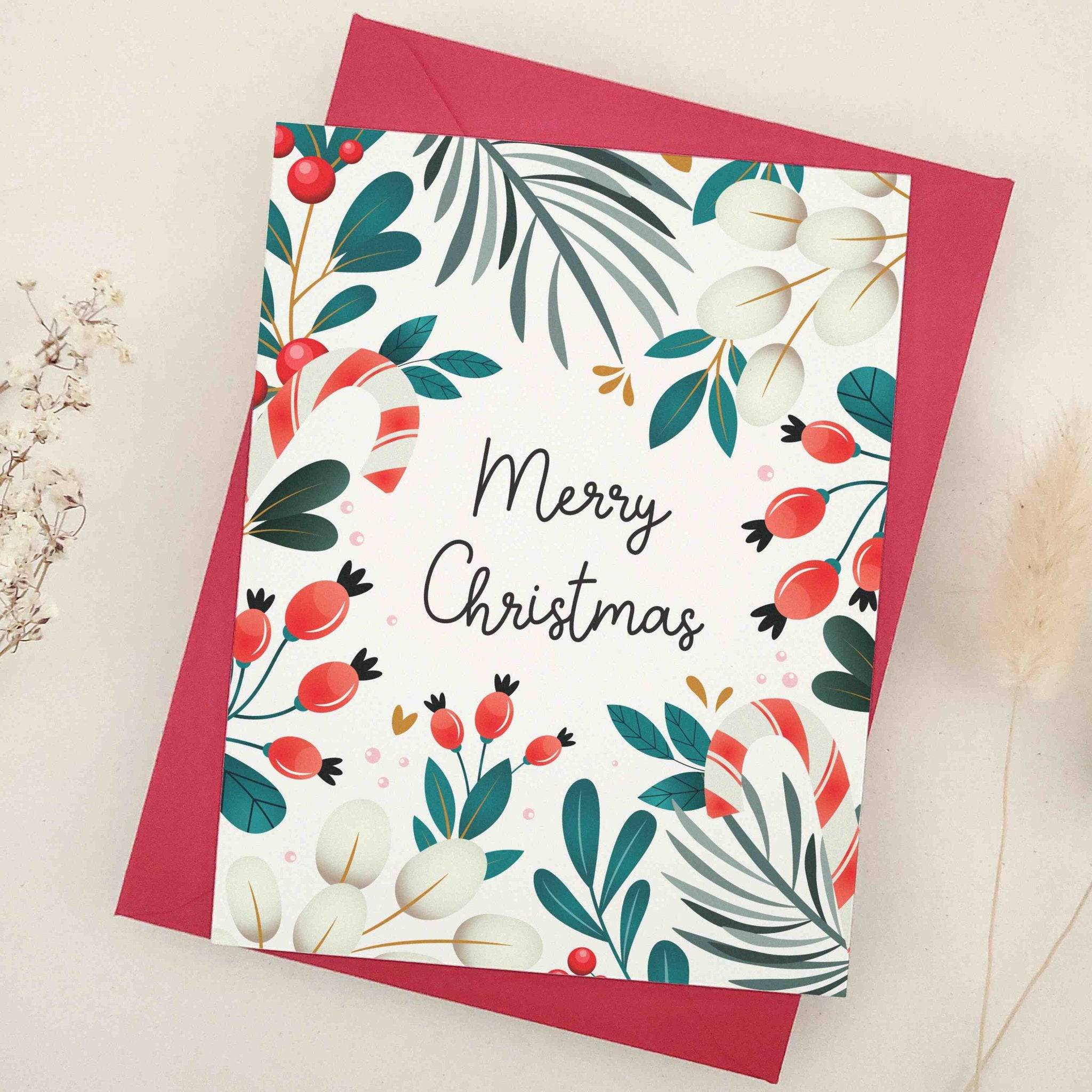 Christmas card on slightly textured off-white cardstock with “Merry Christmas” in black script, surrounded by greenery, red berries, cream leaves, and candy-cane accents, paired with envelope — XOXOKristen