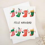 Christmas card with “Feliz Navidad” in bold black text, decorated with a garland of red and green stockings filled with presents, candy canes, and florals, paired with white envelope — XOXOKristen.