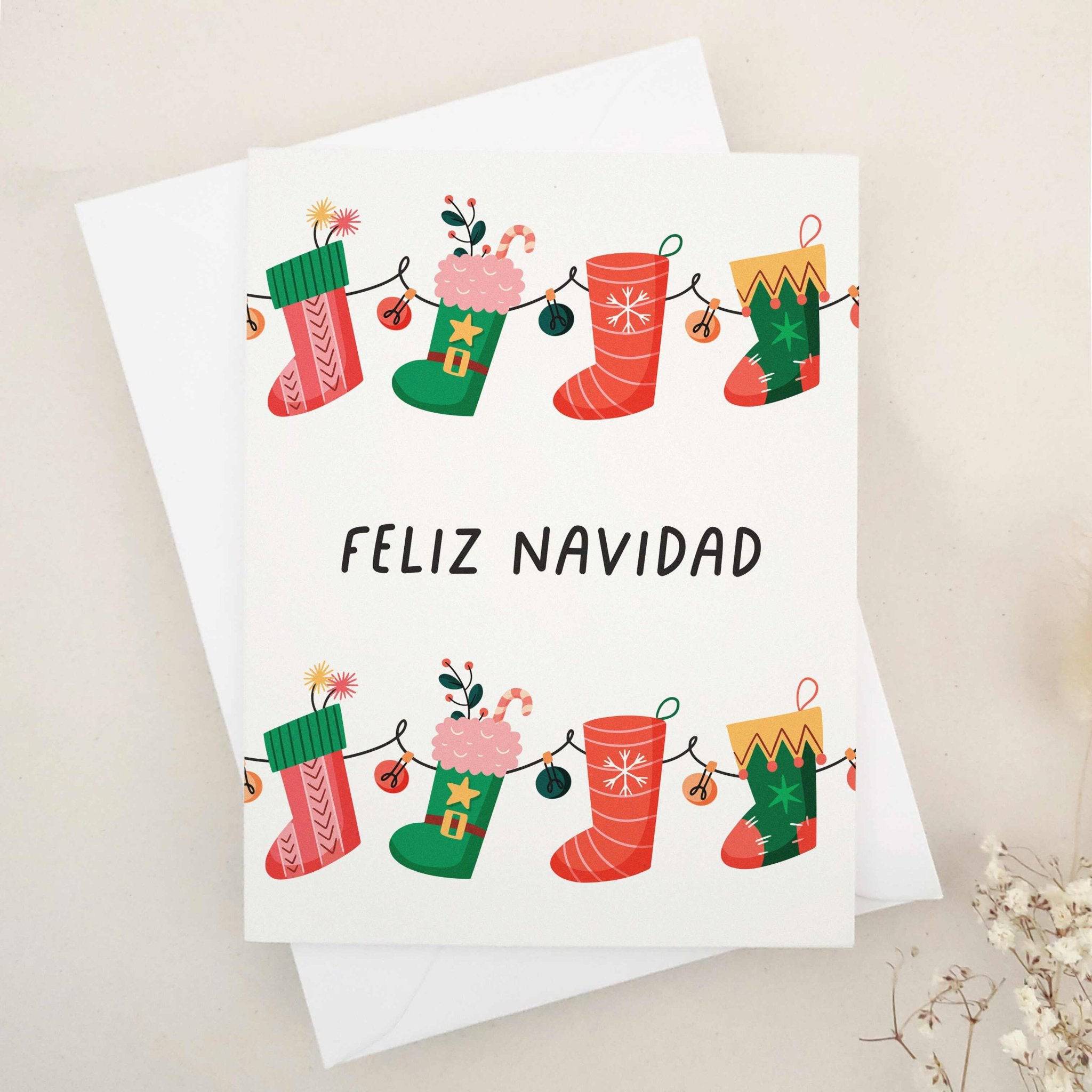 Christmas card with “Feliz Navidad” in bold black text, decorated with a garland of red and green stockings filled with presents, candy canes, and florals, paired with white envelope — XOXOKristen.