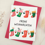 Standing card with festive stocking garland design in red and green, with “Frohe Weihnachten” greeting, styled with red envelope — XOXOKristen.