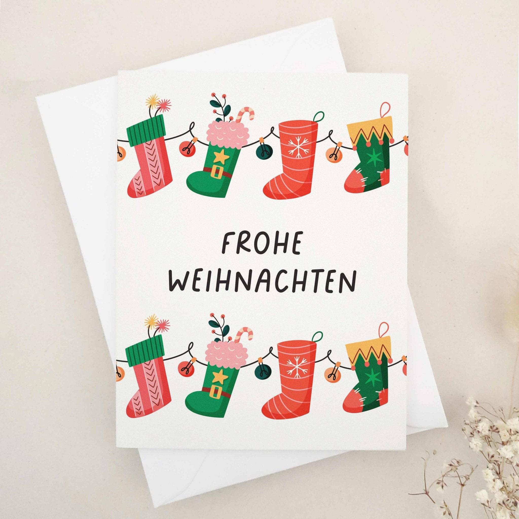 Christmas card with “Frohe Weihnachten” in bold black text, decorated with a garland of red and green stockings filled with gifts, candy canes, and florals, paired with white envelope — XOXOKristen.