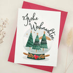 Standing card with festive “Frohe Weihnachten” greeting above snow globe illustration of fir trees, styled with red envelope — XOXOKristen.