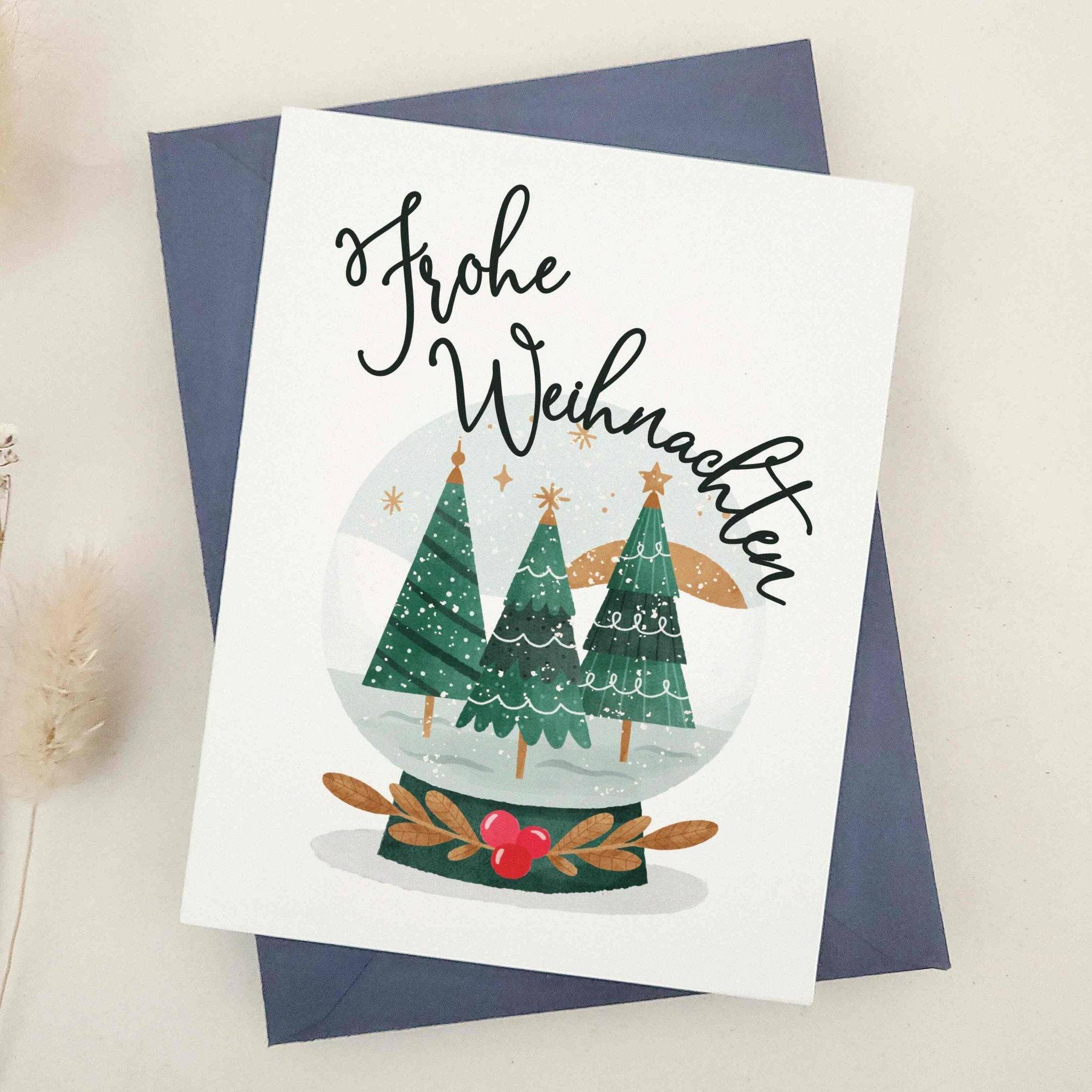 Holiday card with snow globe of green Christmas trees and stars, paired with “Frohe Weihnachten” in script lettering, displayed with blue envelope — XOXOKristen.