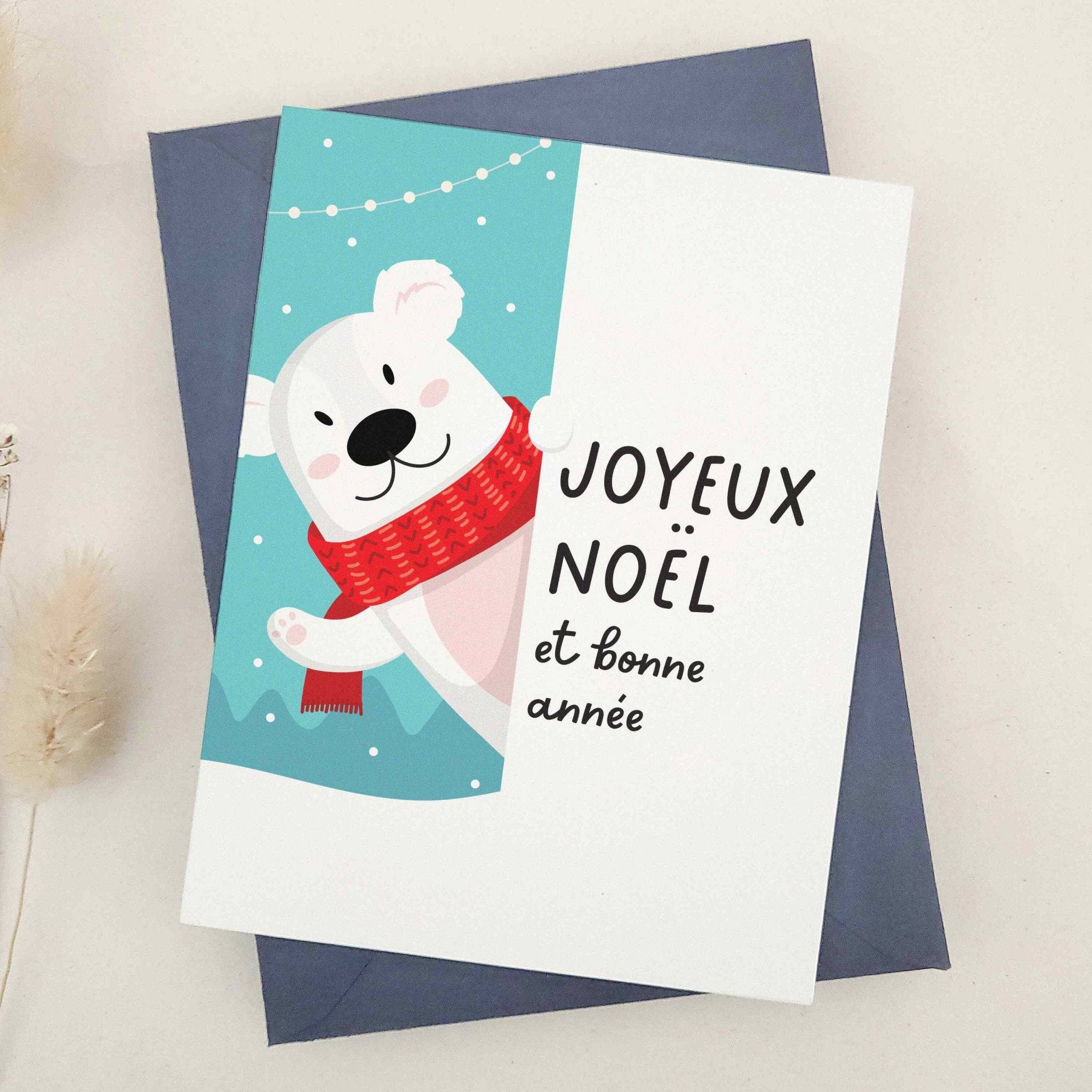 Holiday card featuring polar bear in cozy red scarf on winter blue background with festive lights, paired with “Joyeux Noël et Bonne Année” message, styled with blue envelope — XOXOKristen.