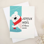 Christmas card with “Joyeux Noël et Bonne Année” in bold black text, illustrated with a smiling polar bear in a red scarf against a snowy blue background with festive string lights, paired with white envelope — XOXOKristen.