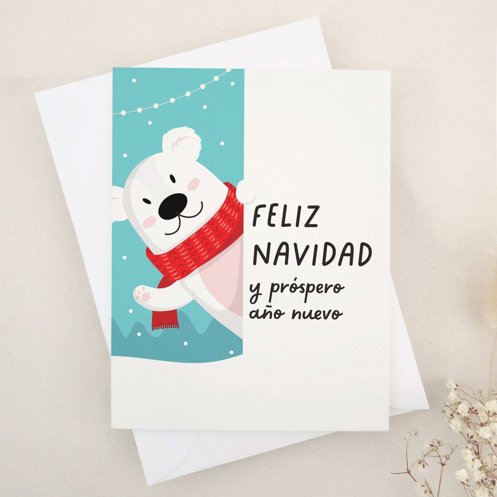 Christmas card with “Feliz Navidad y Próspero Año Nuevo” in bold black text, illustrated with a smiling polar bear in a red scarf against a snowy blue background with festive string lights, paired with white envelope — XOXOKristen.