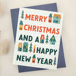 Holiday card featuring bold Christmas message in decorative lettering with scattered snowy details, village houses, and tree icons, styled with blue envelope — XOXOKristen.