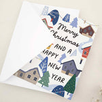 Standing card with festive “Merry Christmas and a Happy New Year” message above a colorful snowy town scene, styled with white envelope — XOXOKristen.