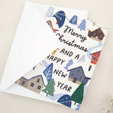 Standing card with festive “Merry Christmas and a Happy New Year” message above a colorful snowy town scene, styled with white envelope — XOXOKristen.