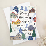 Christmas card with “Merry Christmas and a Happy New Year” in black text, decorated with a colorful winter village of red, yellow, gray, and blue houses, snowy rooftops, and festive trees, paired with white envelope — XOXOKristen.