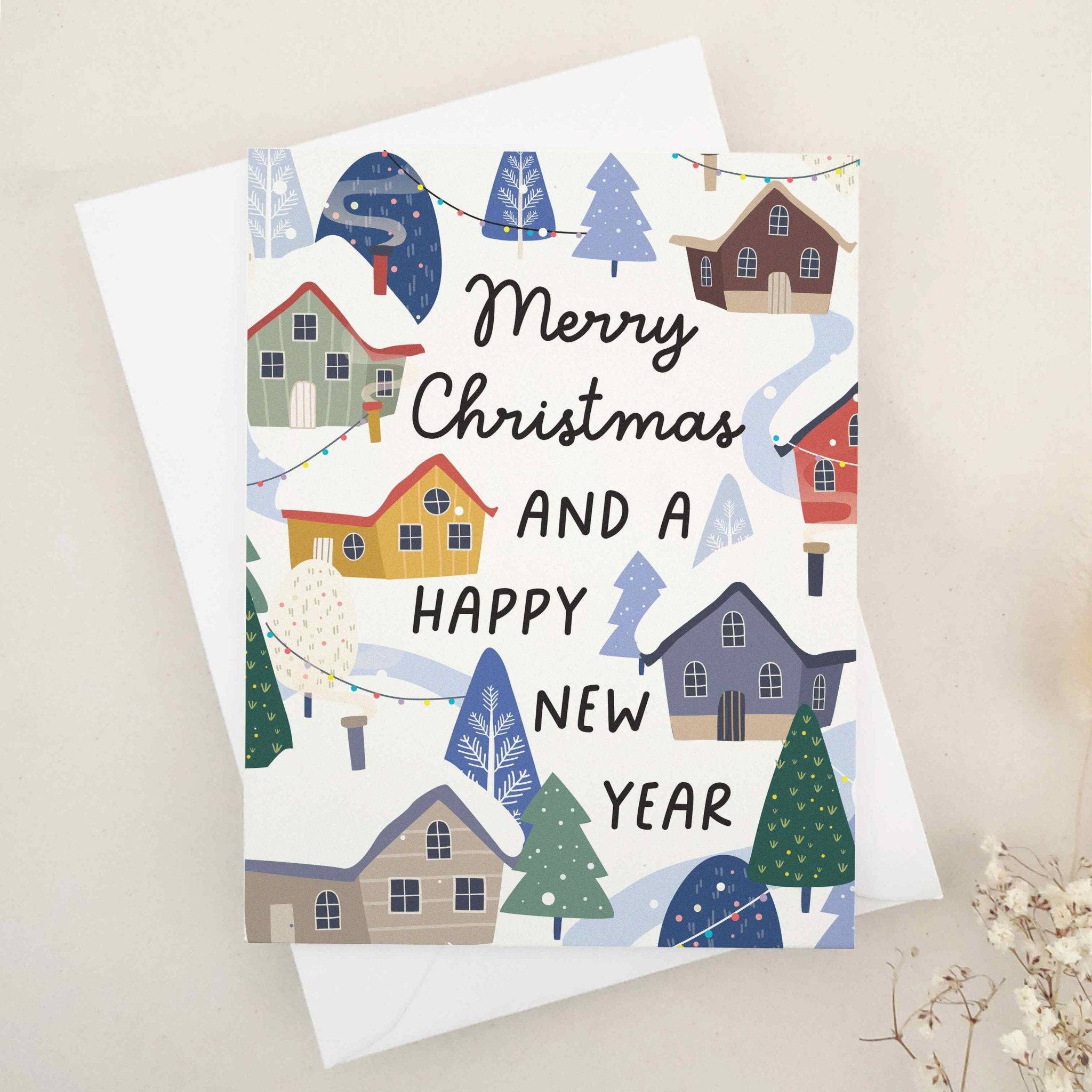Christmas card with “Merry Christmas and a Happy New Year” in black text, decorated with a colorful winter village of red, yellow, gray, and blue houses, snowy rooftops, and festive trees, paired with white envelope — XOXOKristen.