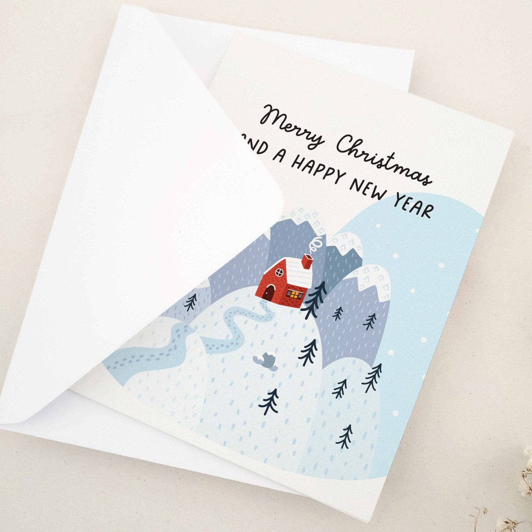 Folded holiday card featuring cozy red cabin among snow-covered mountains and trees with festive greeting, displayed with envelope — XOXOKristen.