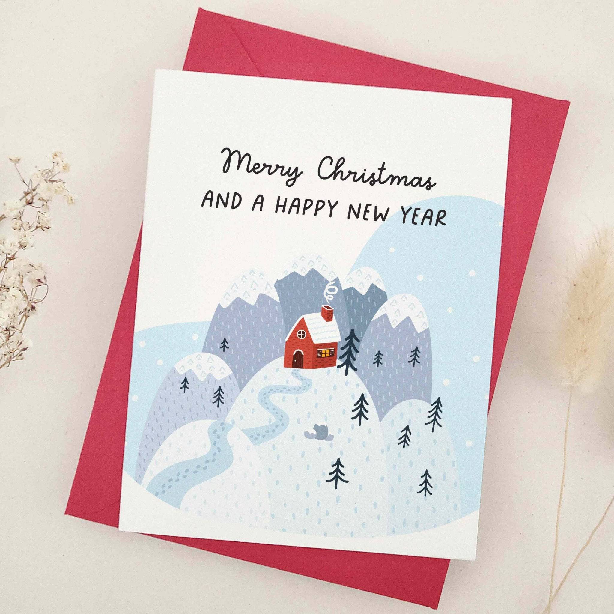 Standing card with “Merry Christmas and a Happy New Year” above a winter mountain scene with winding snowy path and red cabin, styled with red envelope — XOXOKristen.
