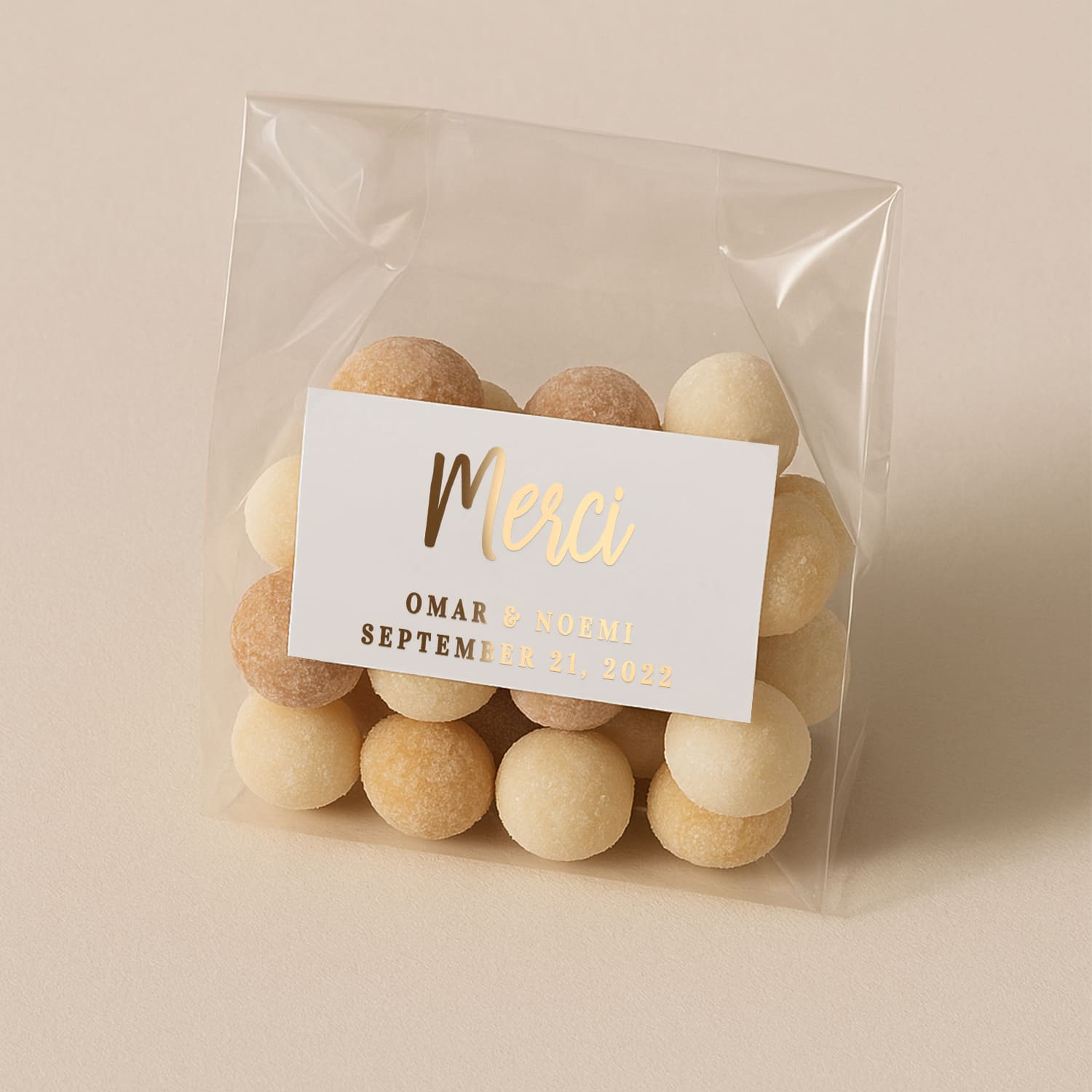 Small personalized white wedding favor sticker with gold foiled Merci text and custom names and date, placed over a small plastic bag with candies - XOXOKristen