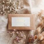 Kraft envelope styled with gold foil rectangular wedding label, handwritten script headline with serif subtext, rustic floral scene – XOXOKristen.