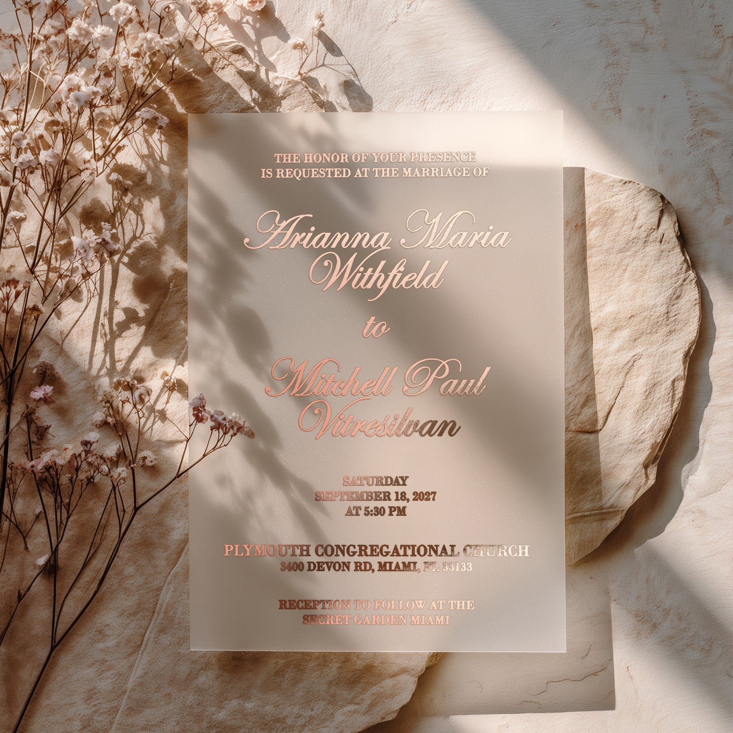 translucent vellum wedding invitation with rose gold foil text, displayed on natural stone surface surrounded by soft floral accents and sunlight, creating a warm, elegant, and romantic mood – XOXOKristen