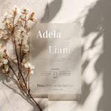 A clear vellum wedding invitation with silver foil text, photographed in natural light beside delicate white cherry blossoms. The soft shadows highlight the translucent texture of the vellum paper, creating a romantic and ethereal look ideal for elegant wedding stationery – XOXOKristen.