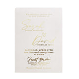 A vertical display of a translucent vellum wedding invitation featuring luminous gold foil lettering, positioned upright among natural stone and dried floral stems. The soft sunlight and gentle shadows enhance the reflective golden accents and the frosted vellum texture, creating a refined, romantic mood. The composition captures the modern luxury of personalized wedding stationery. – XOXOKristen
