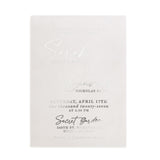 A translucent vellum wedding invitation with metallic silver foil lettering displayed on a natural stone surface beside delicate white blossoming branches. The light and shadows play beautifully across the frosted vellum texture, enhancing the reflective shimmer of the silver foil. The modern fonts and romantic layout create an elegant atmosphere, ideal for spring or garden weddings. – XOXOKristen